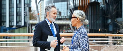 Two businesspeople stand outside and converse while one holds a tablet  