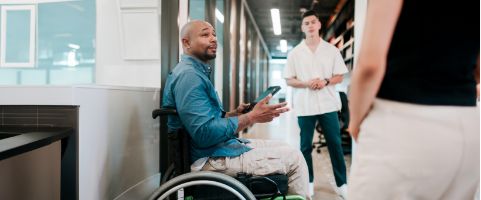 Business professional in wheelchair chatting with 2 colleagues in office building corridor. 