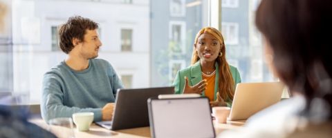 Two people using laptop computers have a discussion at a table.