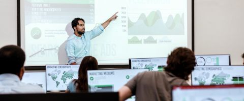 A professor explains a graph displayed on a large classroom screen to students.