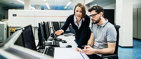 Two people work together in a technology monitoring center. One person with shoulder-length hair, wearing a dark blazer, stands and leans forward with a focused expression, pointing toward a computer screen. The other person, with short hair and glasses and wearing a gray polo shirt, sits at the desk looking at the monitor. A long white desk holds several computer monitors, a keyboard, and a mouse, with large equipment racks visible in the background.
