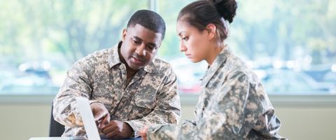 Two people in military uniform look at a laptop while seated at a table.