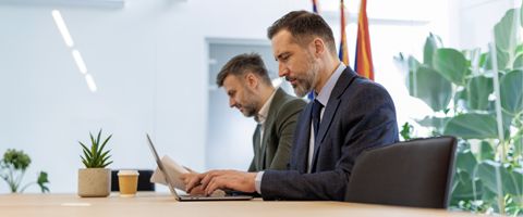Two colleagues sit side by side at a conference table, typing on laptops in a modern office with flags visible in the background.
