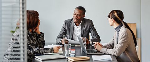Three businesspeople sit at a desk with laptops and notebooks on it and discuss something. 