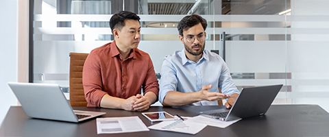 Two businesspeople sit at a desk with two laptops and a tablet and reference something on the screen. 