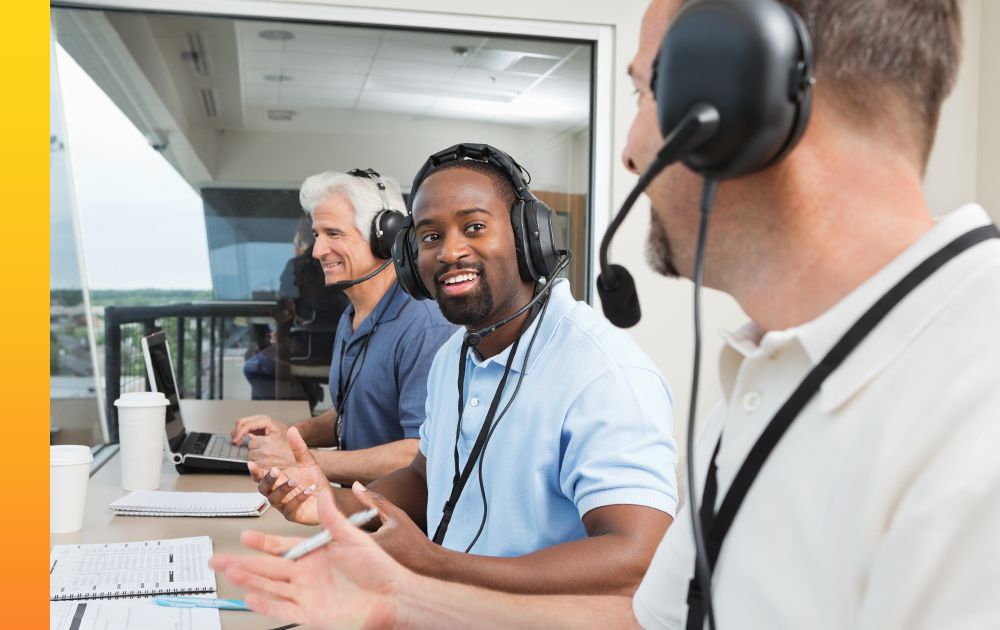 Three commentators with headsets and microphones sit and chat in a studio by a sports field.