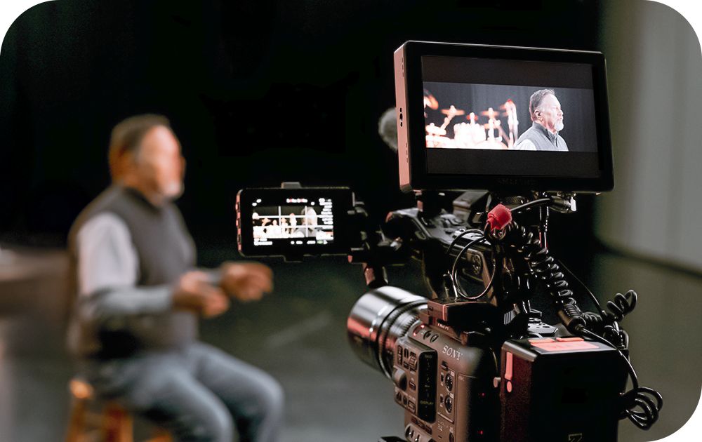 A camera films a person seated on a stage with the subject visible out of focus in the background and clearly framed on the camera’s external monitor.