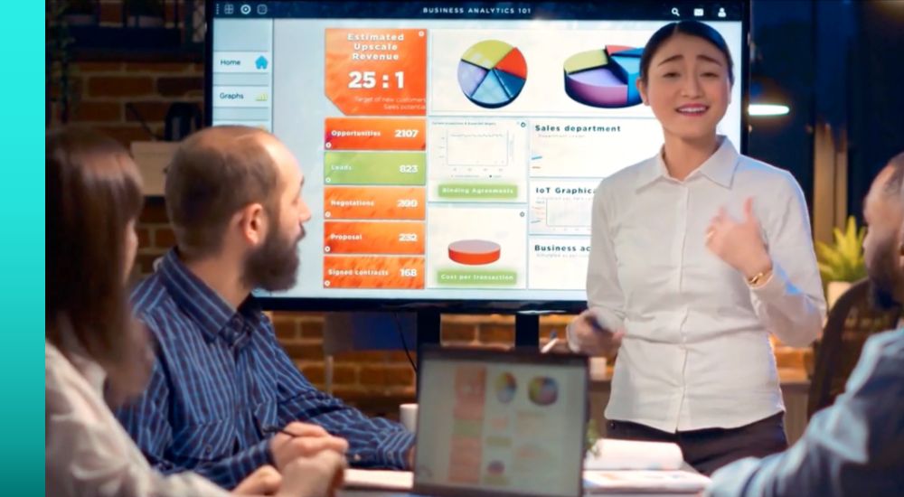 Woman in a white shirt standing and presenting graphs to a group of people at a conference table