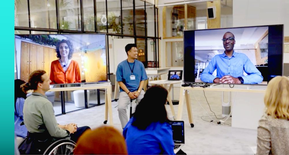  A man sitting on a stool in front of an audience near two large monitors with video call participants