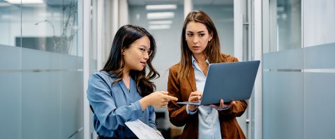 Two colleagues stand in a modern office hallway with glass walls, collaborating over a laptop.