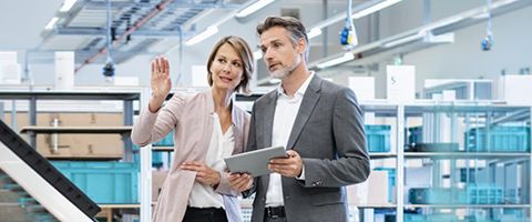 Two people in a warehouse review a tablet while one gestures upward toward shelving.