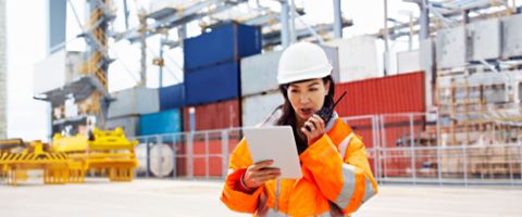 Person in a safety jacket and hard hat reviews a tablet while speaking into a radio at a shipping yard.