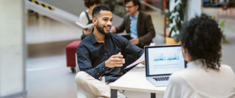 Person gestures while presenting data on a laptop to a colleague in a modern office.