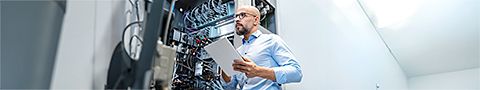 Businessperson in a light blue button-up shirt with glasses and a beard holds a tablet in front of servers