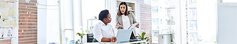 A businessperson in a tan blazer and white button-up stands talking to another businessperson in a white collared shirt sitting at their desk in front of a computer