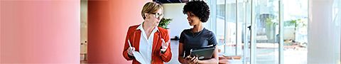 Two businesspeople in a professional setting talk while ones holds a paper and another holds a tablet