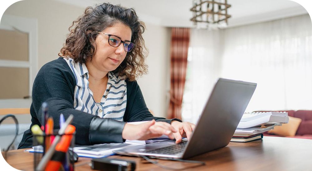 A person with glasses works at a laptop at a table in a living room.
