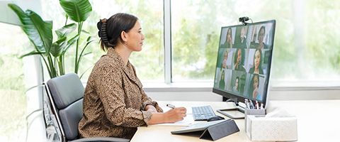 A person sits at a desk and writes while participating in a video call at a desk in front of a computer.