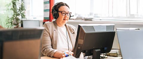 A person in a blazer works at a desktop computer near a window while wearing a headset.