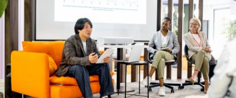 Business presentation with a person sitting on a couch holding a tablet and speaking to a group of seated listeners in a modern office.