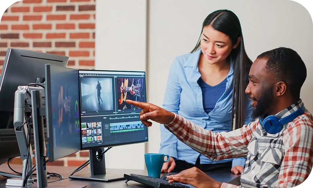 Two coworkers collaborate at a desk, viewing video editing tools and footage on dual computer monitors.