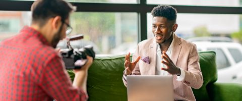 A person in business casual attire talking and holding a laptop on a couch while another person films.