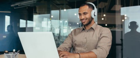 Person in business casual attire sitting in an office setting using a laptop and headphones.