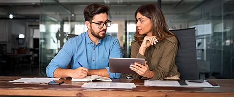 A woman holding a tablet sits next to a man at a desk and discusses the content on the tablet. 