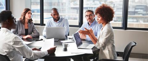 Three people sit at a conference table in front of their laptops and talk to each other