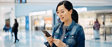 One woman standing in a bright lobby smiles while looking at her cell phone