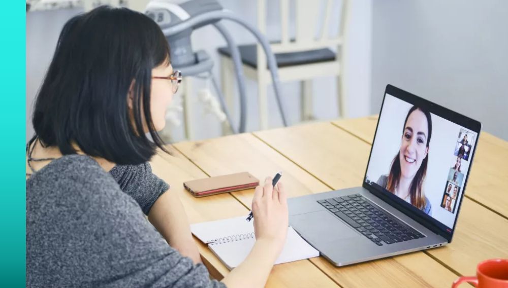 A person sits at a table in front of a notebook and laptop computer and chats with another person on the screen.