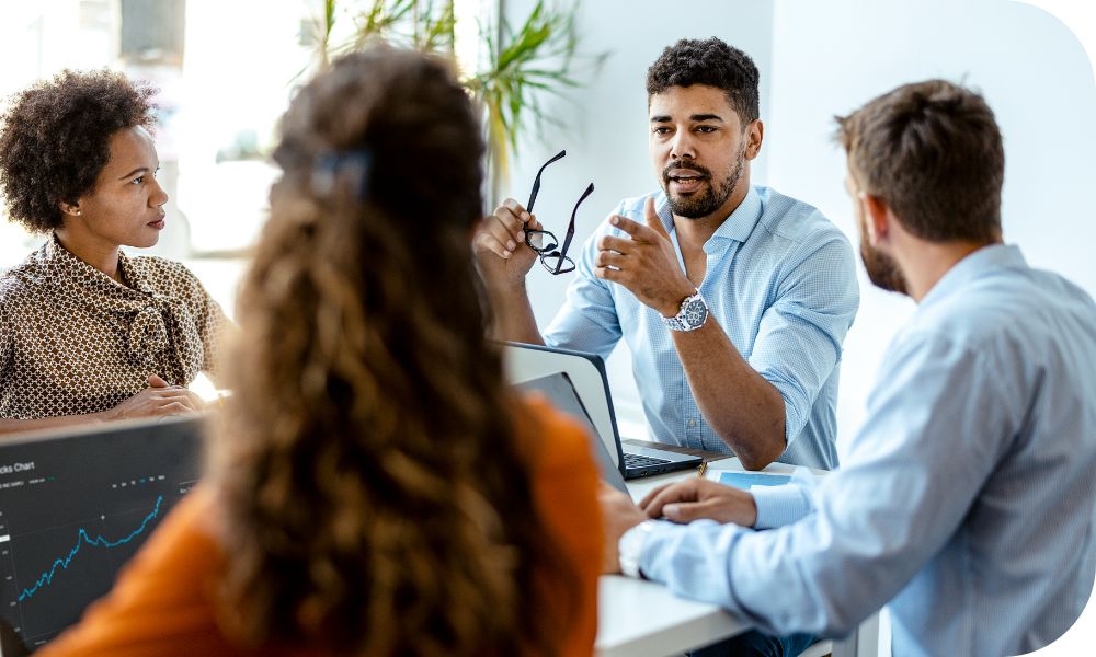 Four businesspeople have a conversation at a desk with laptops in front of them