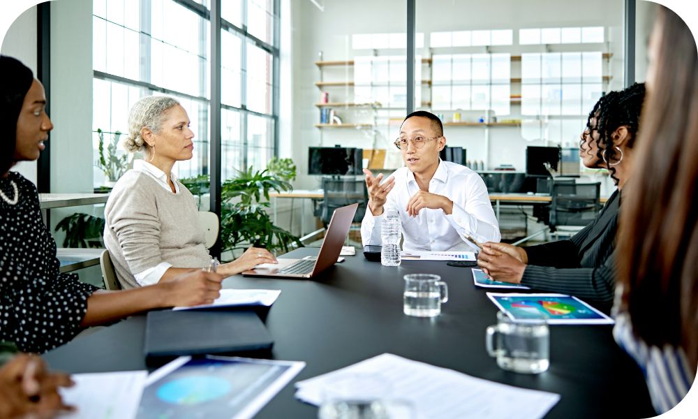 Group of coworkers discussing issues sitting at a conference table with printouts and laptops for reference. 