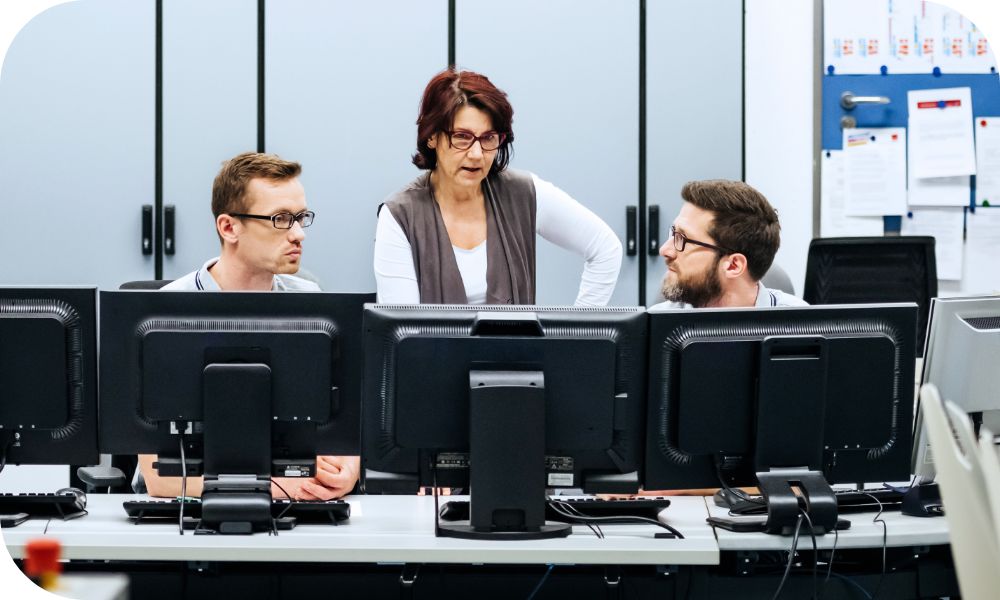 Three businesspeople gathered in front of desktop monitors discussing what’s on their screens