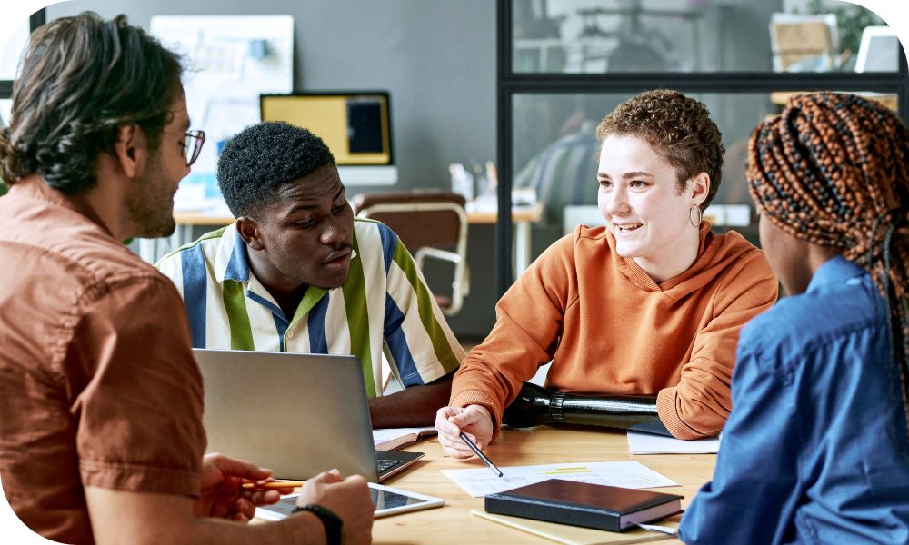 Four businesspeople have a conversation inside an office with a laptop, tablet and a notepad on the desk