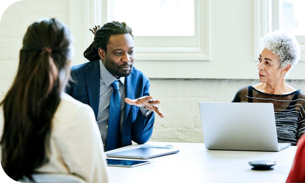 Three people sitting at a conference table with laptops as they watch a man in a blue suit as he is talking