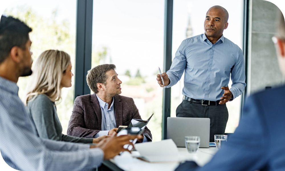 Four businesspeople sitting at a conference table and looking at a fifth person standing and speaking 