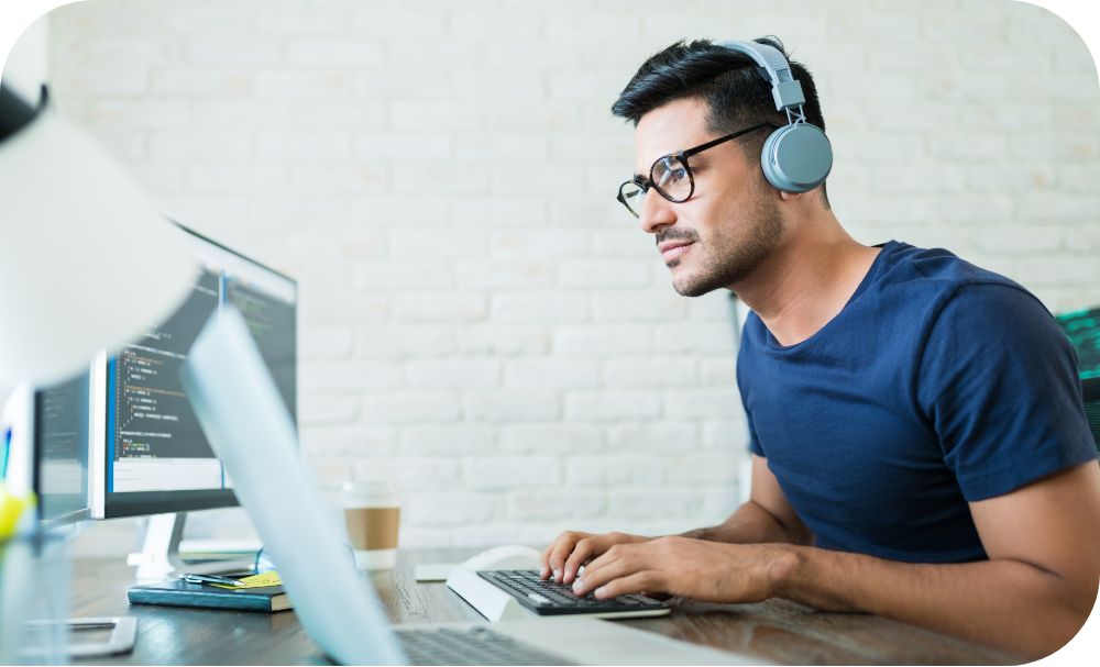 Person wearing headphones and glasses typing on a keyboard in front of computer screens 