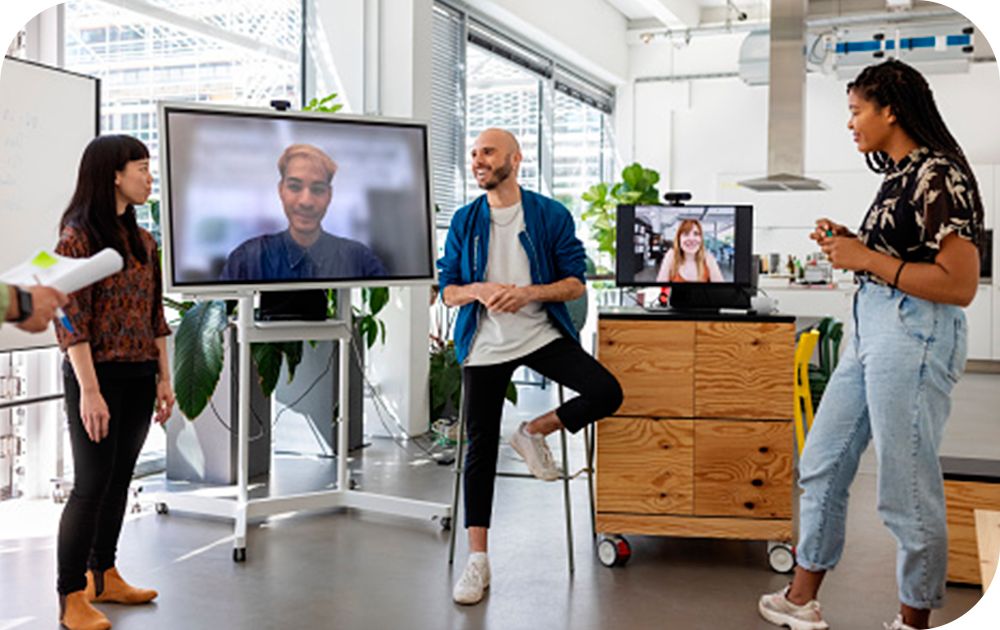 Three people in casual attire standing in an office setting with others on a video call.