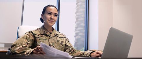 A member of the military wearing camouflage sitting in the office in front of a computer with papers in their hand