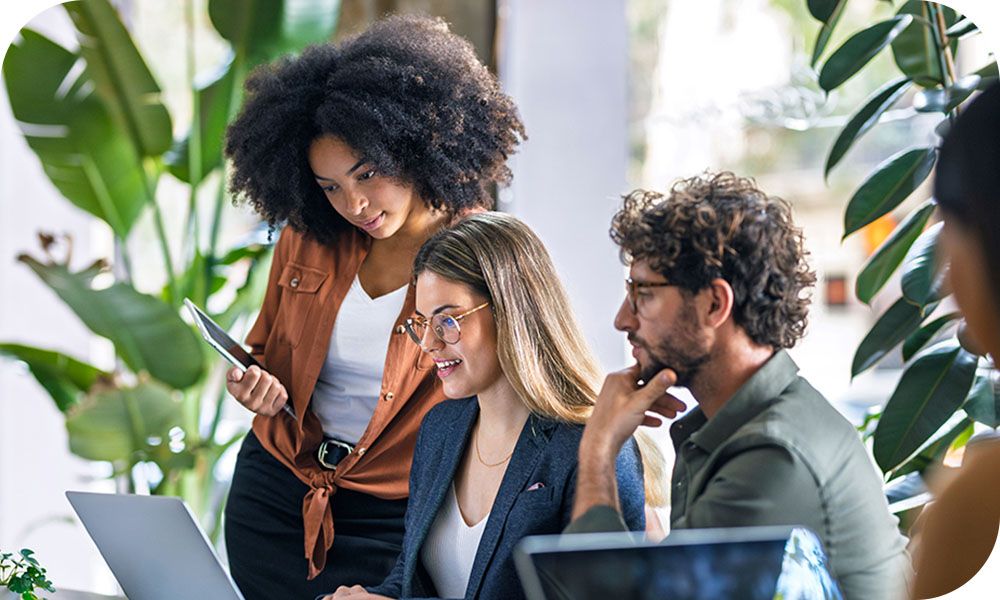 Four coworkers dressed in business casual share a desk and collaborate on laptops.