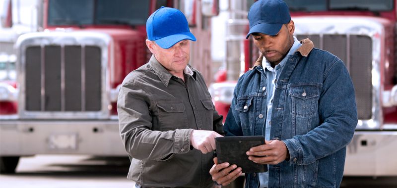 Two men stand in front of trucks and look at a tablet screen