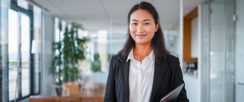 A smiling businessperson in a black blazer and white shirt holds a tablet while standing in a modern office.