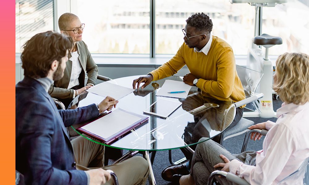 Four people sitting at a round glass table while looking at documents being presented