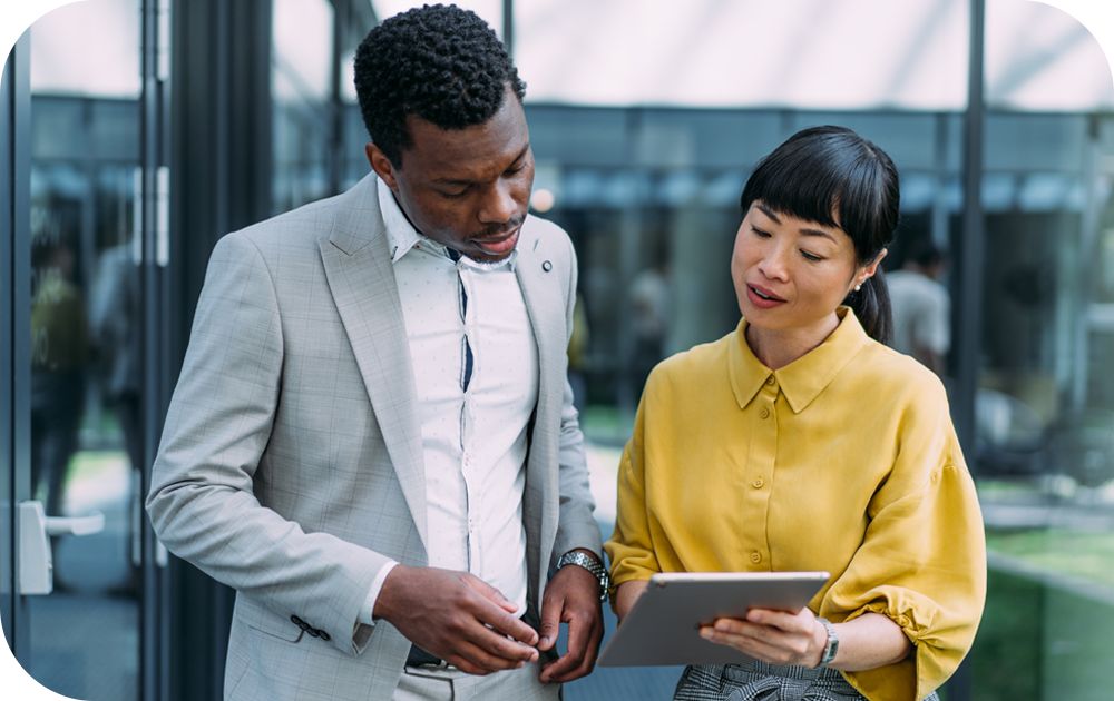 Man in a light gray suit looking down at a tablet device being held by a woman in a yellow shirt