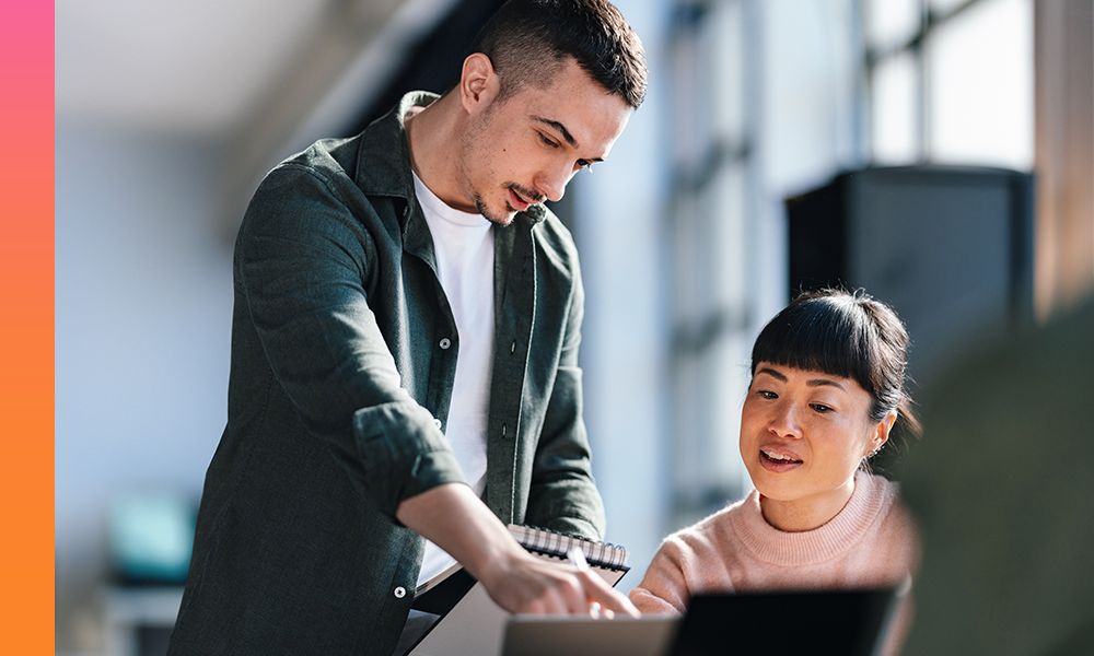 Woman in a pink sweater sitting and working on a laptop while another person standing points to something on her screen