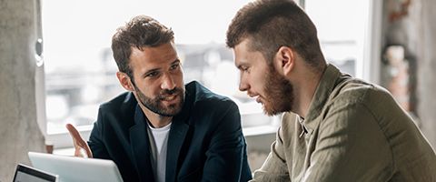 Two men sitting at a table while talking and working on laptops in front of them