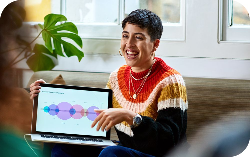 Woman in a multi-color sweater sitting down while smiling and displaying her laptop screen for someone across from her