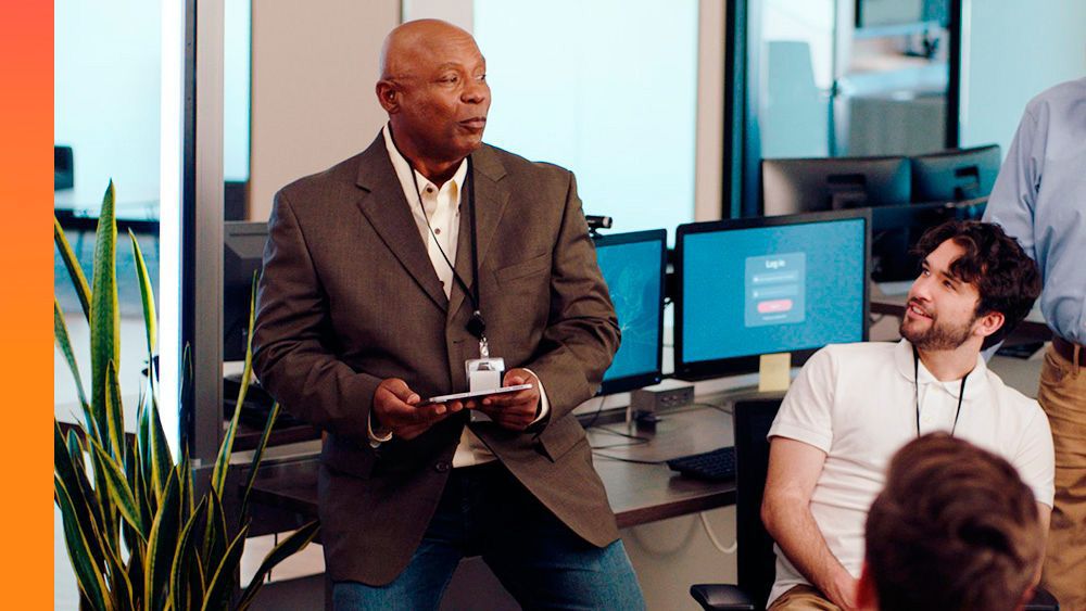 Business meeting in progress, featuring a person in a suit presenting to colleagues in front of multiple computer screens.