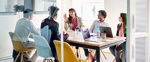 A group of office workers sit around a table in a conference room discussing and gesturing. 